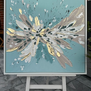 Op de afbeelding: Abstract schilderij op canvas met een witte lijst. Het schilderij heeft een groot, veelkleurig bloemenmotief in tinten teal, wit, goud en bruin. De achtergrond is lichtblauw.
