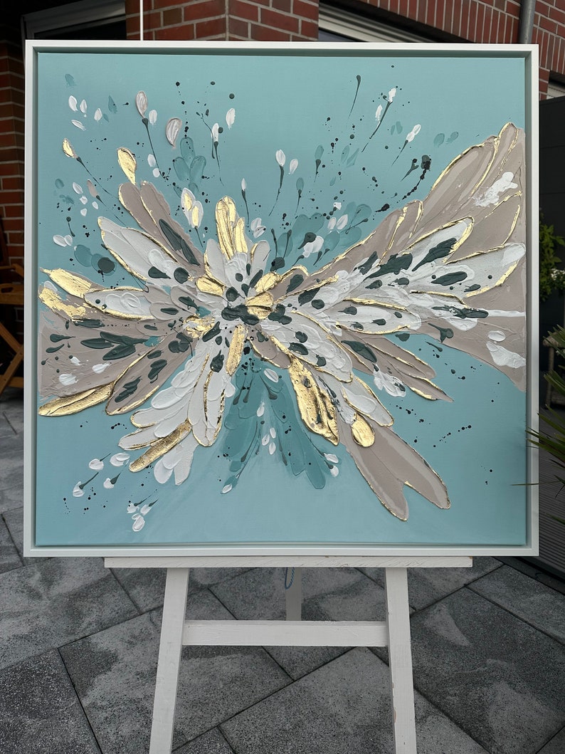 Op de afbeelding: Abstract schilderij op canvas met een witte lijst. Het schilderij heeft een groot, veelkleurig bloemenmotief in tinten teal, wit, goud en bruin. De achtergrond is lichtblauw.