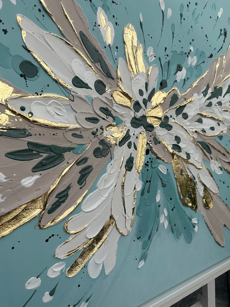 Op de afbeelding: Abstract schilderij met een groot, gestileerd bloemmotief in tinten wit, goud en turquoise op een lichtblauwe achtergrond.