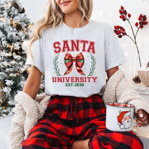 May include: A light gray t-shirt with the text "SANTA UNIVERSITY EST.1836" in red and green. The shirt features a red bow and laurel wreath design. The person is wearing red and black plaid pajama pants and holding a mug with a Santa Claus graphic.