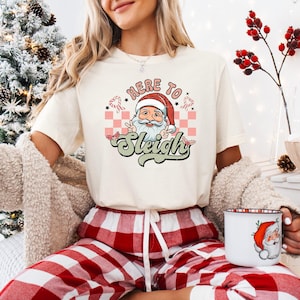 May include: Cream-colored t-shirt with a Santa Claus graphic and the text "Here to Sleigh". The Santa graphic is in a circle with a pink and white checkered background. The person is wearing red and white plaid pajama pants.