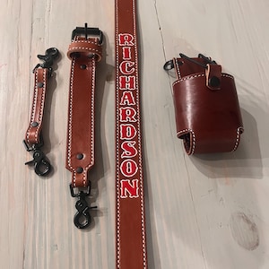May include: A brown leather strap with the name "RICHARDSON" embroidered in red. The strap has two black metal clips and a brown leather pouch with a black metal clip.