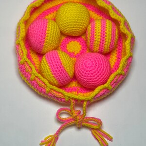 May include: A handmade crochet set featuring a round basket and five balls. The basket and balls are in bright pink and yellow. The basket has a scalloped edge and a drawstring closure with a bow. The balls are either solid yellow or pink and yellow striped.