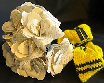 Puede incluir: Un ramo de rosas de papel hechas con las páginas de un libro, con un lazo de ganchillo amarillo y negro. Las páginas del libro son de color crema y el texto es visible. El lazo está hecho de hilo y tiene un patrón a rayas.