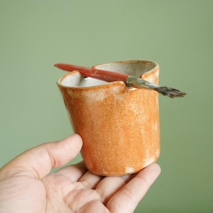 May include: A small, handmade ceramic cup in shades of orange and white, with a speckled, rustic texture. A red-handled paintbrush rests across the top, suggesting its use for art or crafts.