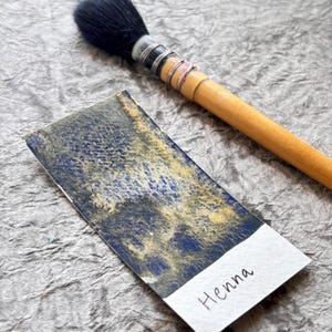 May include: A watercolor swatch with blue and gold hues, labeled "Henna," next to a wooden-handled brush with a black bristle head. The swatch is on a textured, gray surface.