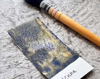 May include: A watercolor swatch with blue and gold hues, labeled "Henna," next to a wooden-handled brush with a black bristle head. The swatch is on a textured, gray surface.