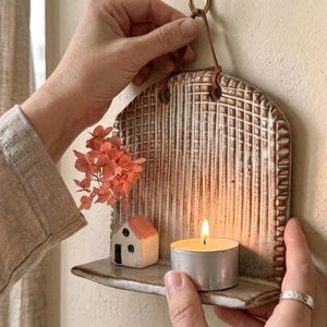 Può includere: Una mensola da parete in ceramica con un pannello posteriore strutturato a motivo grigliato. La mensola presenta una piccola statuetta di casa in ceramica, un rametto di ortensia rosa essiccata e un lumino acceso sulla superficie.