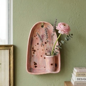 May include: A wall-mounted ceramic vessel with an arched backplate in a mottled pink and brown glaze. A small cylindrical pocket holds a single pink ranunculus flower and two sprigs of dried lavender, secured by a small brass-colored screw.