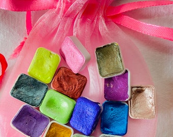 May include: A collection of watercolor paint pans in a variety of colors, including green, blue, purple, gold, and silver. The pans are arranged in a circle and are sitting in a pink organza bag.
