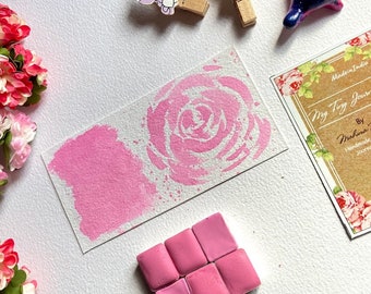 Può includere: Una collezione di acquerelli rosa e un disegno di rosa ad acquerello su una carta bianca. La carta presenta un campione di vernice rosa e un disegno di rosa. L'immagine include anche un biglietto da visita e clip decorative a forma di unicorno.