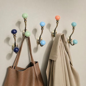 Könnte beinhalten: Sechs Wandhaken mit Messing-Finish. Jeder Haken hat einen farbigen Keramikknopf in Blau, Grün und Orange. Eine braune Ledertasche und ein beiger Mantel hängen an den Haken.