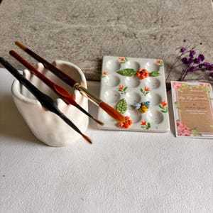 May include: A white ceramic paint palette with 12 wells, decorated with orange flowers, green leaves, and a bee. The palette is next to a white ceramic brush holder with four paintbrushes.
