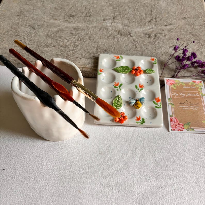 May include: A white ceramic paint palette with 12 wells, decorated with orange flowers, green leaves, and a bee. The palette is next to a white ceramic brush holder with four paintbrushes.