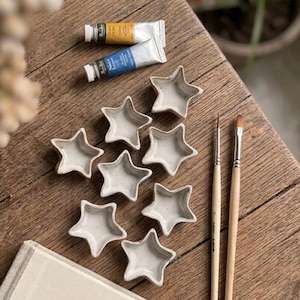 Peut inclure: Une collection de petits plats en céramique en forme d'étoile, de couleur neutre, disposés sur une surface en bois. Deux tubes de peinture et deux pinceaux sont également présents. L'arrangement suggère un thème artistique ou artisanal.