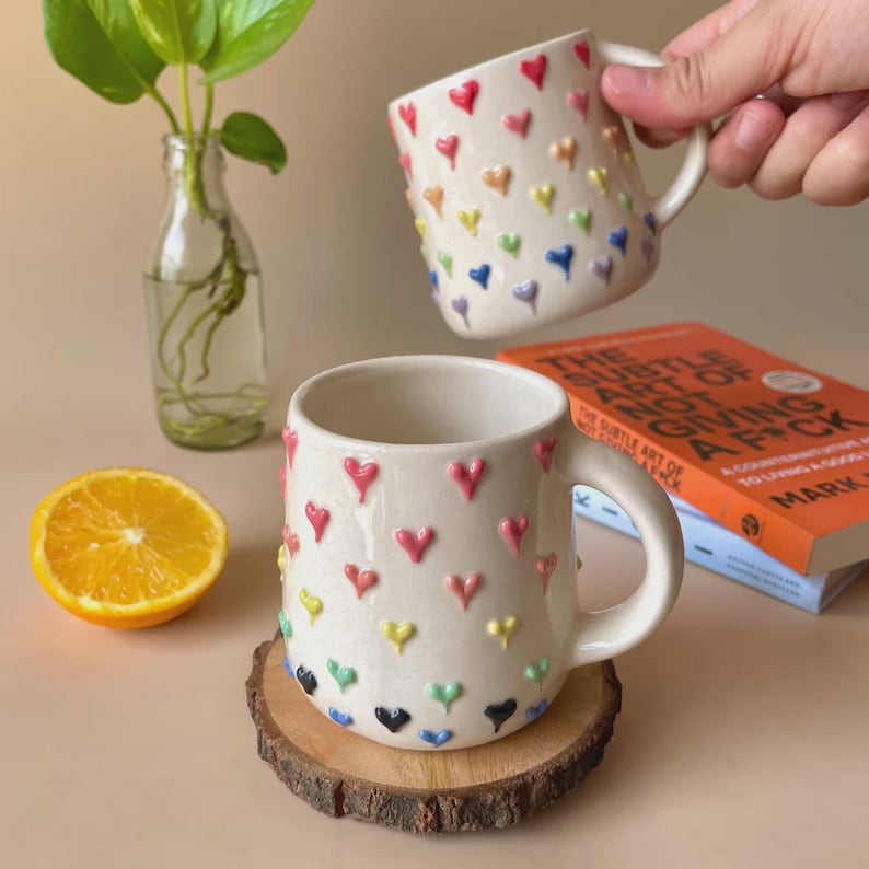 May include: Two white ceramic mugs with raised, colorful heart designs. One mug is on a wooden coaster, and the other is held by a hand. The mugs have a handle and are filled with a liquid. A sliced orange and a book are also in the image.