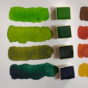 May include: Watercolor paint swatches in shades of green, from light to dark, alongside their solid paint block counterparts. The swatches are painted on white paper, showcasing the color variations.