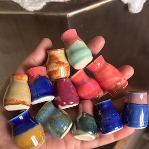 Op de afbeelding: Een verzameling van twaalf miniatuur keramische vazen in diverse kleuren, waaronder blauw, roze, geel, groen en rood. Elk klein vaasje heeft een gladde, geglazuurde afwerking met opvallende kleurverlopen en gespikkelde texturen op de ronde vormen.