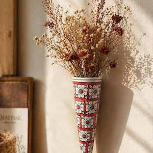 Könnte beinhalten: Konisch geformte Wandvase mit rotem und weißem Blumendesign, gefüllt mit Trockenblumen. Die Vase hat ein sich wiederholendes Muster aus weißen Blumen auf rotem Grund. Die Trockenblumen sind braun, beige und burgunderrot.