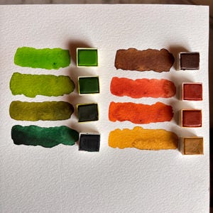 May include: Watercolor paint swatches in shades of green, brown, orange, and yellow. Each color is displayed as a rectangular swatch and in a small square pan. The swatches are on white paper.