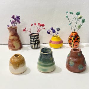 Op de afbeelding: Een verzameling van zeven kleine, handgemaakte keramische vazen in verschillende vormen, kleuren en patronen. Sommige vazen bevatten gedroogde bloemen. Kleuren zijn bruin, geel, groen en oranje. De vazen staan op een witte ondergrond.