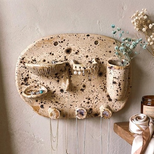 Può includere: Un organizer da parete ovale in ceramica con finitura maculata beige e marrone. Presenta due piccole ciotole per anelli, una barra orizzontale con fori per orecchini, un vaso cilindrico alto e tre ganci inferiori per le collane.