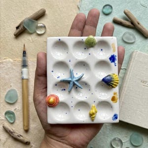 Op de afbeelding: Een wit keramisch kunstenaarspalet met 12 putjes, versierd met blauwe verfspatten, miniatuur schelpen en een blauwe zeester. Een aquarelkwast en een klein boek zijn ook zichtbaar.