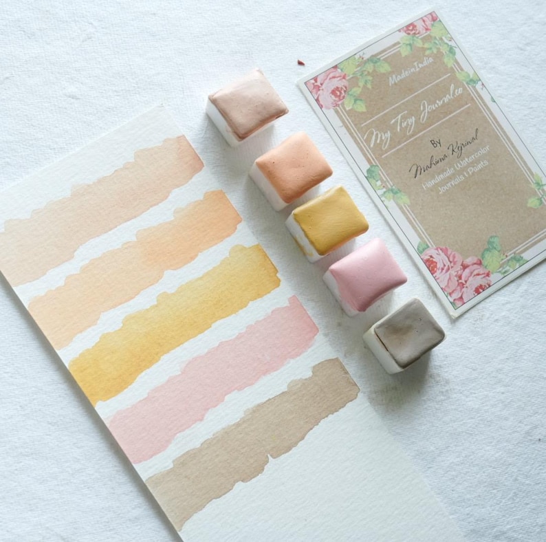 May include: Watercolor paint set with six square pans in shades of beige, peach, yellow, pink, and brown. The colors are displayed on a white paper with matching swatches. A card with floral accents and text that reads "My Tiny Journalie" is also present.