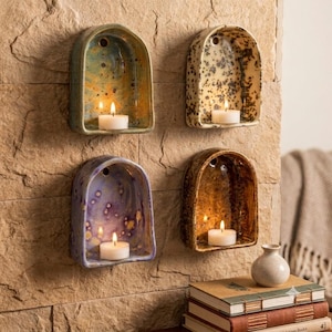 May include: Four arched ceramic wall sconces mounted on a stone surface, each holding a white tea light candle. The sconces feature distinct glazed finishes: one mottled blue and yellow, one cream with black speckles, one purple with light spots, and one brown with a glossy texture.