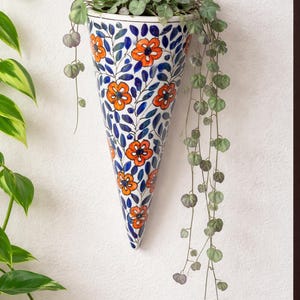 Op de afbeelding: Een kegelvormige keramische wandplantenbak met een witte basis en een handgeschilderd patroon van feloranje bloemen en donkerblauwe bladeren. De plantenbak is gevuld met een hangende groene plant met kleine hartvormige blaadjes.