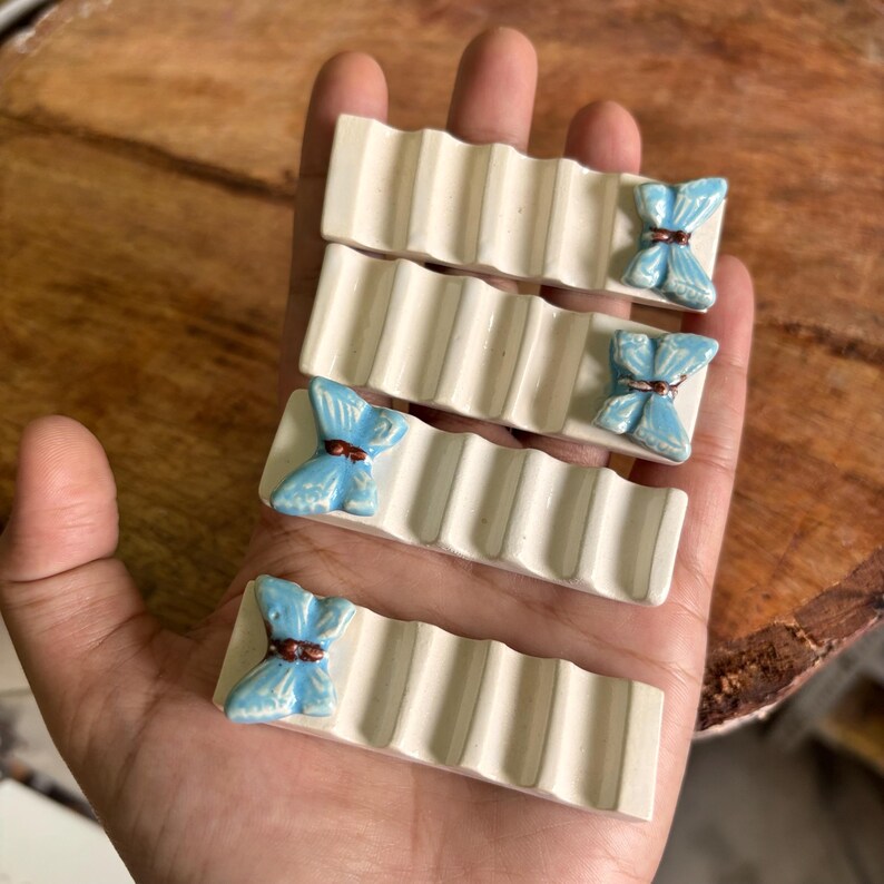 Puede incluir: Cuatro reposa palillos de cer&aacute;mica blanquecina con un dise&ntilde;o acanalado. Cada reposa palillos presenta un acento de mariposa azul claro con un centro marr&oacute;n. Los reposa palillos se sostienen en una mano.