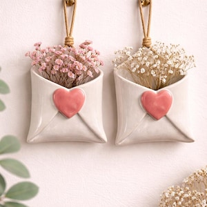 May include: Two white ceramic wall-mounted envelope-shaped planters, each featuring a raised pink heart on the front. The planters are suspended by tan rope loops and contain dried flowers, with pink baby's breath in one and white in the other.
