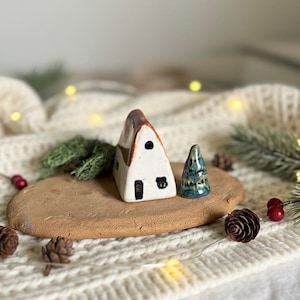 May include: A ceramic miniature house with a brown roof and white walls, next to a small blue and green ceramic Christmas tree. The house and tree sit on a brown, oval-shaped base, with pinecones and greenery around them.