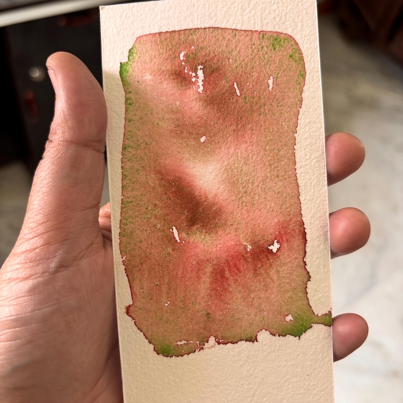 May include: A watercolor swatch on a rectangular piece of paper. The colors are primarily red and brown, with green accents along the edges. The paper is held by a hand.