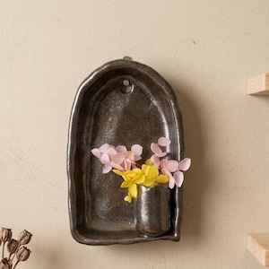 Può includere: Un vaso da parete in ceramica a forma di arco, di colore marrone scuro con una finitura lucida e maculata. L'oggetto presenta una piccola tasca cilindrica integrata alla base che contiene un mazzetto di delicati fiori secchi rosa e gialli.