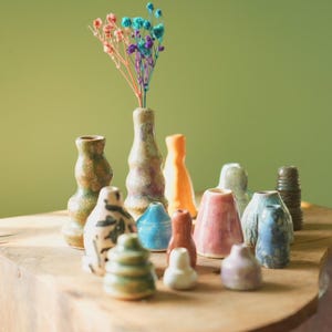 Könnte beinhalten: Eine Sammlung kleiner, handgefertigter Keramikvasen in verschiedenen Formen und Farben, darunter Grün, Blau, Orange und Rosa. Einige Vasen haben strukturierte Oberflächen. Eine Vase enthält bunte Trockenblumen. Die Vasen werden auf einer Holzoberfläche ausgestellt.
