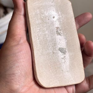 Pu&ograve; includere: Un ciondolo rettangolare in ceramica bianco sporco con una superficie leggermente strutturata. Il ciondolo ha bordi arrotondati e sembra presentare alcune imperfezioni. Il ciondolo &egrave; tenuto in mano, suggerendo che si tratti di un oggetto indossabile o di un materiale artigianale.