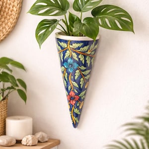 Peut inclure: Un pot mural bleu en forme de cône avec un motif floral, contenant une plante verte à grandes feuilles. Le pot a un bord blanc et est décoré de fleurs et de vignes bleues, oranges et jaunes. Le pot est fixé sur un mur blanc.