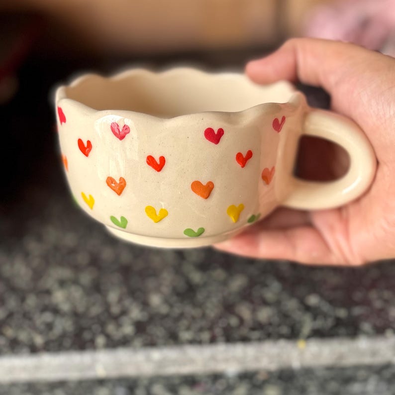 May include: A cream-colored ceramic mug with a scalloped rim and a handle. The mug is decorated with colorful heart shapes in red, orange, yellow, and green. The mug is held in a hand.