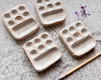 Puede incluir: Cuatro paletas de pintura de cerámica con un esmalte blanco roto moteado y un borde natural sin esmaltar. Cada paleta tiene diez pozos circulares y un área de mezcla rectangular. Un pincel con mango de madera y cerdas oscuras descansa cerca.