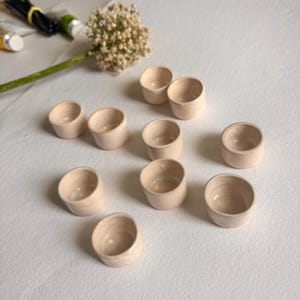 May include: Ten small, cream-colored ceramic bowls with a slightly textured surface. The bowls are arranged in a semi-circle on a white surface.