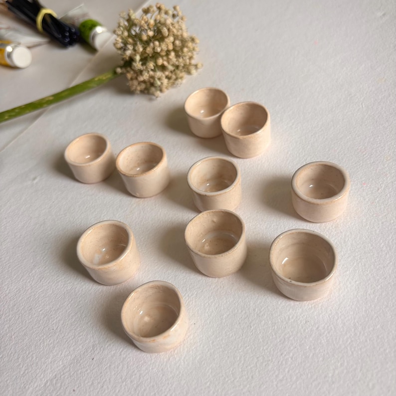 May include: Ten small, cream-colored ceramic bowls with a slightly textured surface. The bowls are arranged in a semi-circle on a white surface.