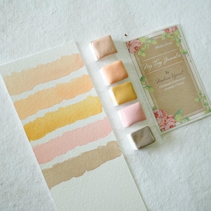 May include: Watercolor paint set with a range of warm colors, including peach, yellow, and pink. A watercolor swatch card displays the colors. The set includes a card with the text "My Tiny Journaler" and floral accents.