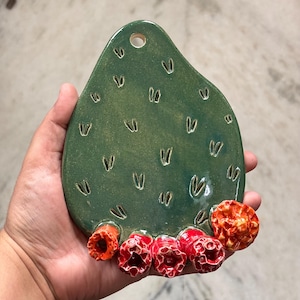 May include: A teardrop-shaped green ceramic wall hanging featuring a glossy finish with small incised V-shaped markings across the surface. At the bottom, five textured, bright red and orange flower-like protrusions are attached to the base.