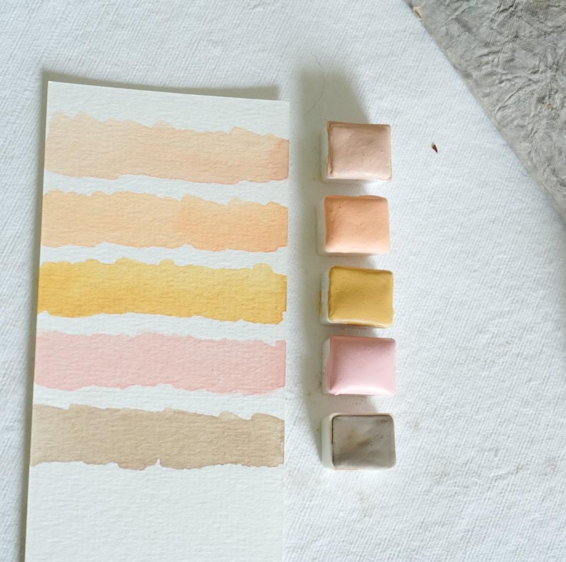 May include: A watercolor paint swatch card with six horizontal color bands in shades of peach, yellow, pink, and brown. Five square watercolor paint pans in matching colors are arranged vertically to the right.
