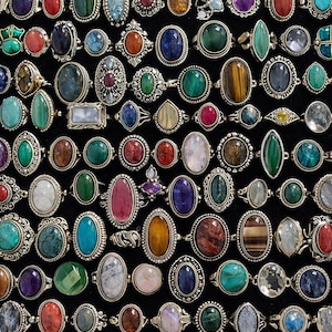 Può includere: Una vasta collezione di anelli assortiti con varie pietre preziose colorate e cabochon incastonati in fasce di metallo color argento. Gli anelli presentano un'ampia gamma di forme, tra cui ovali, rotonde, rettangolari e a navetta.