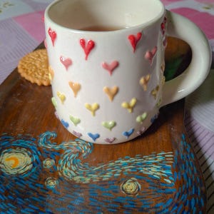 May include: A white ceramic mug adorned with colorful heart-shaped embellishments, filled with tea, rests on a wooden tray. The tray features a painted design inspired by Van Gogh's "Starry Night", with a biscuit on the side.