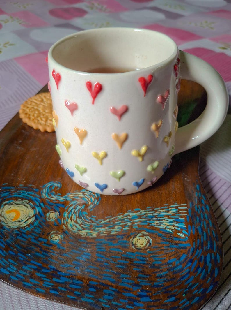 May include: A white ceramic mug adorned with colorful heart-shaped embellishments, filled with tea, rests on a wooden tray. The tray features a painted design inspired by Van Gogh's "Starry Night", with a biscuit on the side.