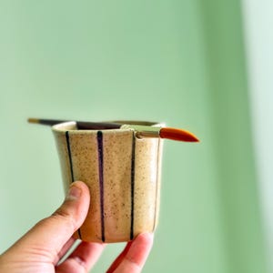 Op de afbeelding: Een gespikkelde beige keramieken beker met verticale donkerbruine strepen en een inkeping in de rand. Een penseel met een donkere steel en een feloranje punt ligt horizontaal over de beker, rustend in de inkeping van de rand.