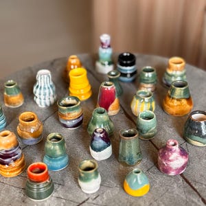 Può includere: Una collezione di piccoli vasi in ceramica fatti a mano in varie forme e colori. I vasi presentano una gamma di smalti, tra cui blu, verde, giallo e marrone. Alcuni hanno superfici strutturate, altri sono lisci. I vasi sono disposti su una superficie di legno.